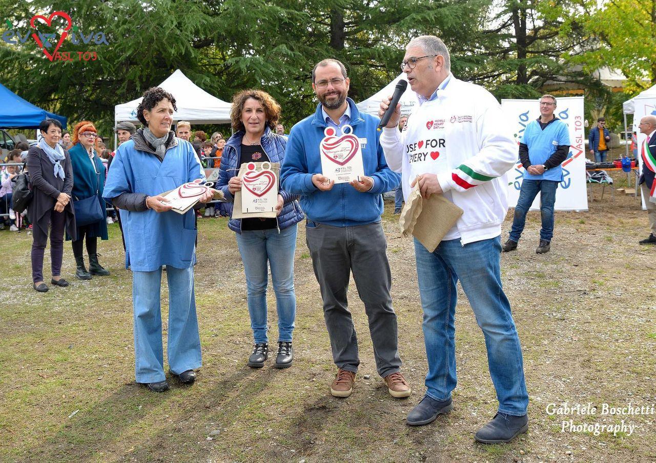 Outdoor award ceremony: four people holding heart-shaped plaques, one speaking into a microphone